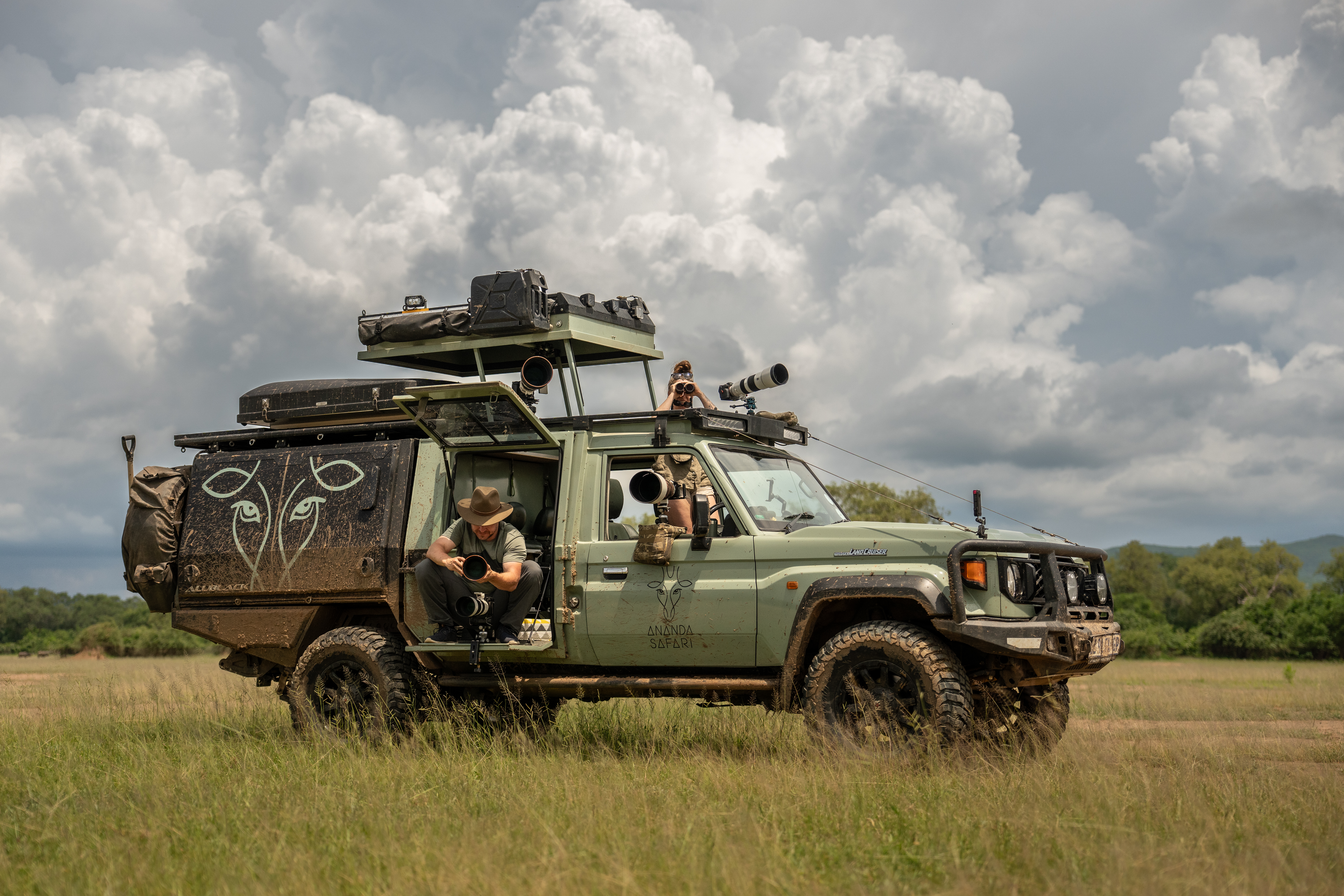 An Ananda Safari game-viewing vehicle, kitted out with roof tents and gear, parked on open grassland beneath a wide, cloud-filled sky